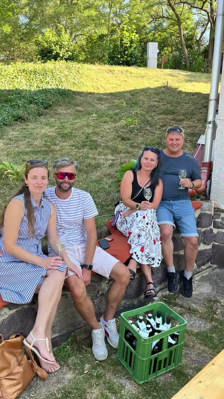 Hosté se sklenkami vína sedí na zídce před sklepem